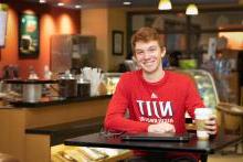 College student sitting in a coffee shop with a coffee cup and his laptop smiling wearing a NJIT shirt